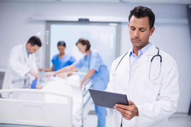 Doctor standing in hospital room using digital tablet while medical team attends to patient in background. Ideal for illustrating modern healthcare, medical technology, and professional medical environments. Useful for healthcare websites, medical articles, and technology in medicine promotions.