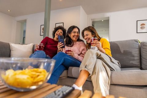 Diverse women laughing on sofa with drinks and snacks, cozy friends night in at home