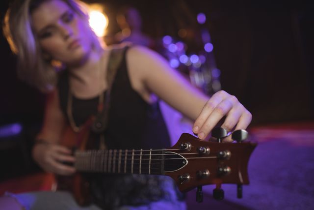 Female musician adjusting the tuning peg of her guitar on stage in a nightclub. Ideal for use in articles about live music, concert promotions, nightlife events, and musician profiles.