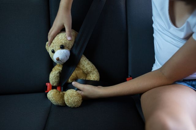 Teenage girl securing teddy bear with seatbelt in car back seat. Ideal for themes on child safety, travel, and childhood care. Can be used in articles, advertisements, and educational materials promoting vehicle safety and child protection.