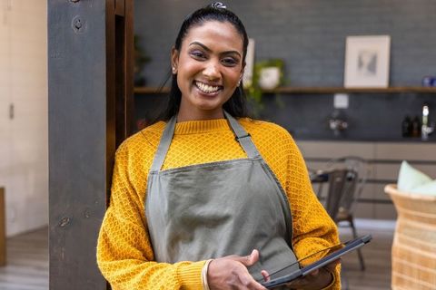 Smiling indian barista leaning on post holding tablet in cozy modern cafe interior
