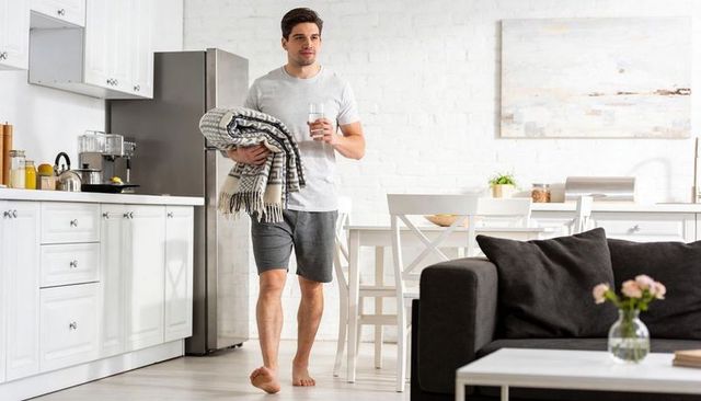 Young man walking carrying folded throw and glass of water in bright modern kitchen