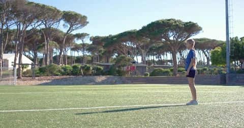 Youth female soccer player standing on sunlit turf with long shadow and pine trees
