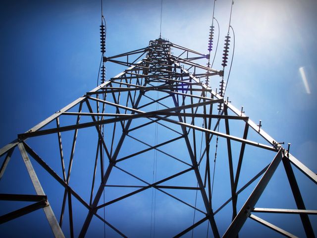Showing a transmission tower from a low angle against a blue sky. Suitable for themes like energy industry, power infrastructure, technology, and construction. Can be used for educational purposes, industry reports, and marketing in energy or construction sectors.