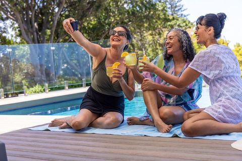 Diverse senior women taking selfie and toasting cocktails poolside on sunny deck