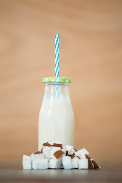 Coconut smoothie in a glass bottle with a green polka dot lid and blue striped straw, surrounded by chopped coconut pieces on a wooden surface. Ideal for use in health and wellness blogs, tropical drink recipes, vegan lifestyle promotions, and organic product advertisements.