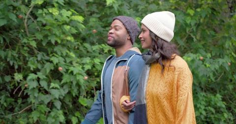 Diverse couple walking autumn trail wearing knit beanies mustard-yellow sweater blue jacket