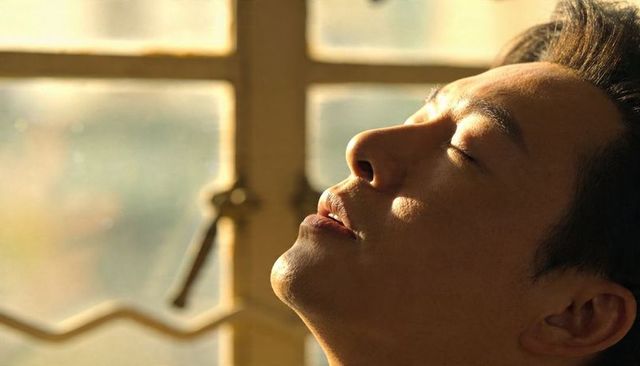 Asian man basking in golden hour sunlight, eyes closed, relaxed profile by window
