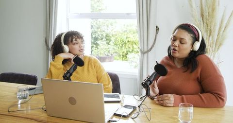 Female co-hosts recording conversational podcast from home studio with laptop, microphones
