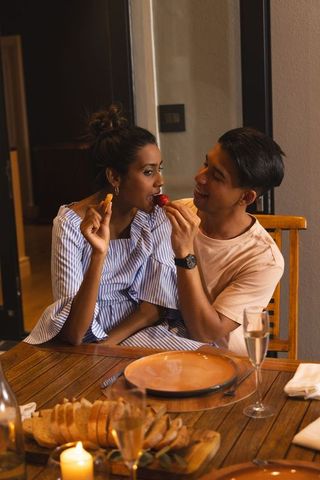 Couple sharing strawberry during candlelit dinner, feeding romantic bite at wooden table