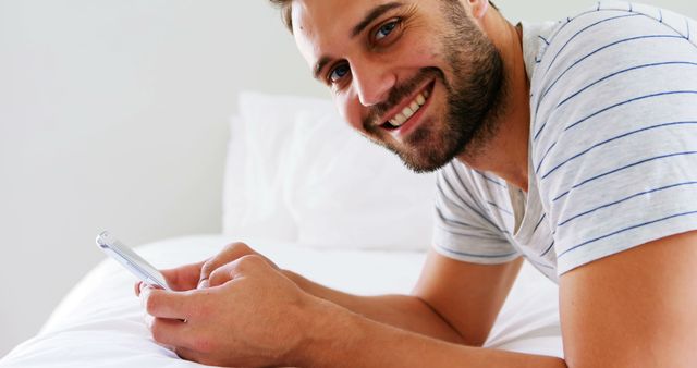 Young man lies on bed with white sheets, holding and using smartphone. Man is smiling and appears relaxed in casual striped T-shirt. Ideal for promoting technology, lifestyles, leisure, and comfort.