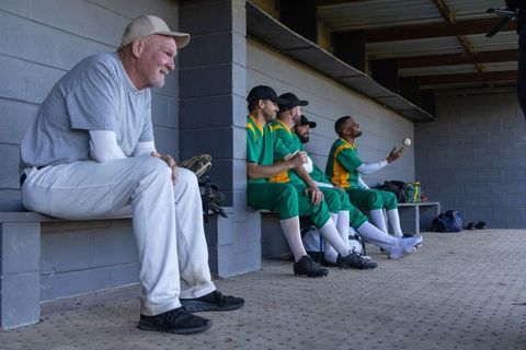 Veteran coach watching team in dugout while players tossing ball, relaxing, preparing for game