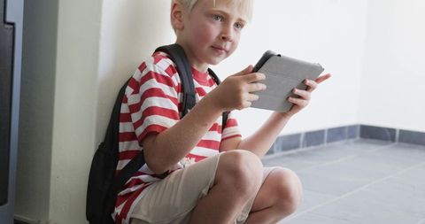 Blond boy crouching in hallway using tablet with backpack ready for school learning