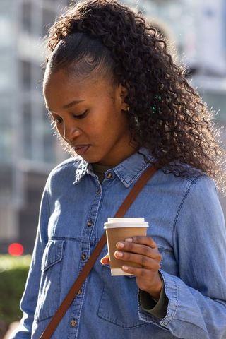 African american woman walking through urban plaza holding coffee cup in sunlit denim shirt