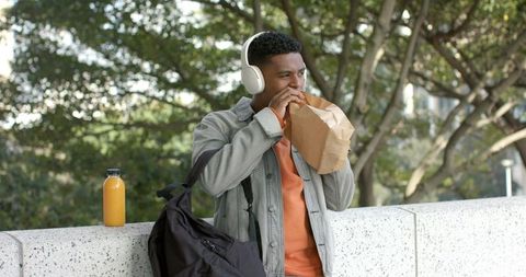 African american student enjoying lunch with headphones on campus ledge
