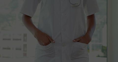 Doctor standing in white lab coat with hands in pockets by bright clinic window stethoscope