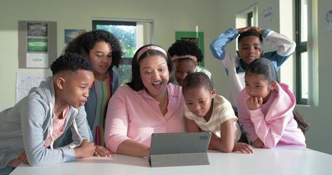 African American teacher with elementary students gathering around tablet, reacting with joy