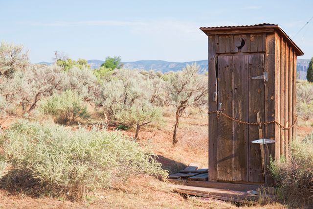 Rustic wooden outhouse stands in an arid desert landscape amid sparse vegetation and clear skies. This old-fashioned structure offers a glimpse into rural living. This image is suitable for use in articles, blogs, and advertisements centered around countryside life, outdoor adventures, sustainable living, and rural experiences.