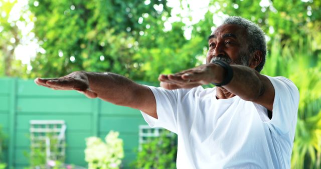 This image shows an elderly man practicing yoga stretches outside on a sunny day, surrounded by lush greenery. He is wearing a white shirt and appears focused and calm as he extends his arms in front of him. Ideal for articles and promotions on senior fitness, health and wellness programs, meditation apps, or outdoor activity advertisements targeted towards older adults.