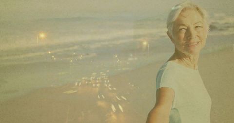 Smiling senior woman reaching hand toward camera by seaside with dusk light trails