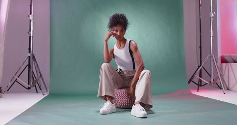 African american model posing on knitted pouf sitting in minimalist studio green backdrop