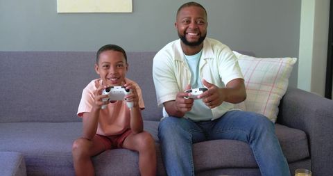 African american father and teenage son playing video games on couch smiling together