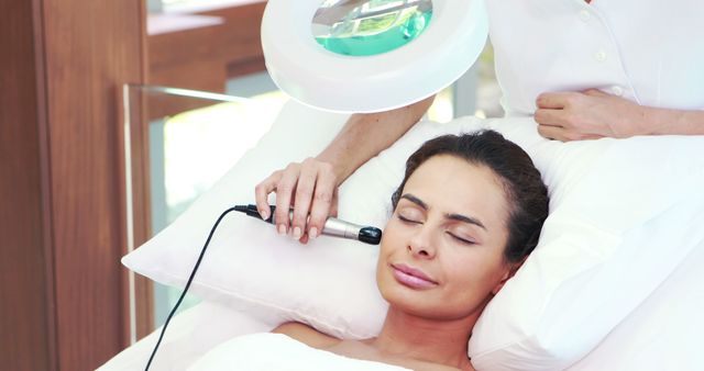 Woman receiving facial skin treatment in a spa, lying comfortably with eyes closed as professional uses device on her face. Ideal for promoting spa services, beauty treatment advertisements, self-care rituals, and relaxation imagery.