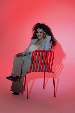 Confident african american woman posing on red chair in blazer and heels on pink gradient