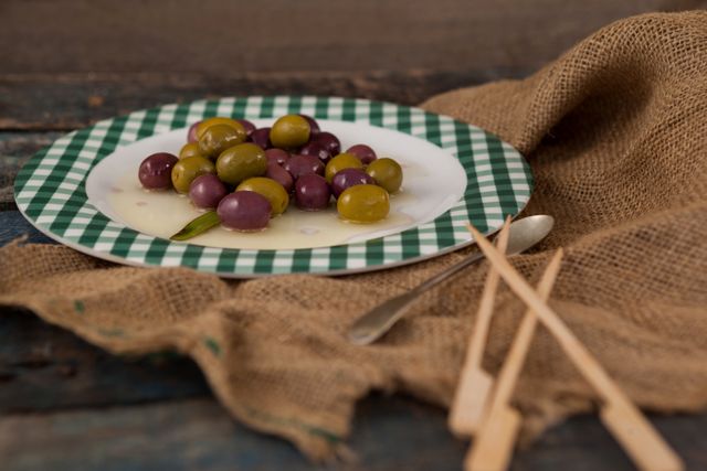 Close up of green and black olives served on a plate with a rustic wooden table background. Burlap cloth and wooden utensils add to the rustic feel. Ideal for use in food blogs, Mediterranean cuisine promotions, healthy eating campaigns, and restaurant menus.