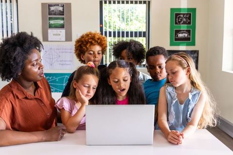 Teacher guiding diverse students gathering around laptop for collaborative learning