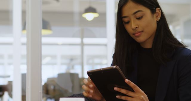 Young biracial woman reviews data on a tablet in an office setting. Her focused expression suggests she's analyzing important information for work.