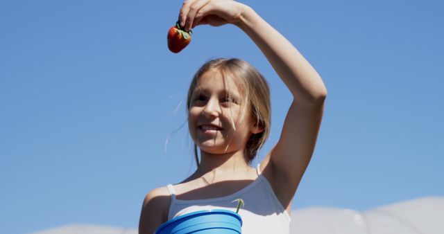 A young girl enjoys a sunny day at a farm, holding a fresh strawberry she picked. This image is perfect for use in promotions related to outdoor activities, farming, agricultural projects, healthy foods, and children’s activities. It conveys joy, warmth, and the fulfillment of harvesting fresh produce.