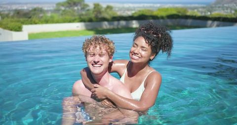 Laughing and hugging diverse couple in infinity pool with coastal ocean view luxury getaway
