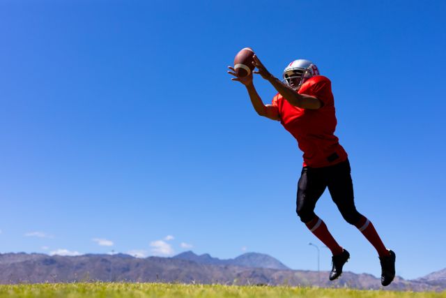 This image depicts an African American football player in mid-air, jumping to catch a ball during a game on a sunny day. The background features clear blue skies and distant mountains. Ideal for use in sports-related articles, athletic training materials, or promotional content for football events.