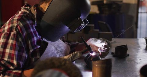Welder using TIG torch and filler rod on metal pipe at workshop bench, protective gear