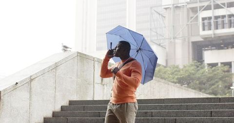 African American man walking stairs holding blue umbrella talking on smartphone in rain