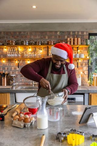 African american man wearing santa hat whisking batter in holiday kitchen with string lights