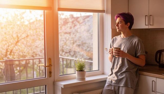 Golden hour morning coffee by balcony, young man holding mug in cozy kitchen apartment