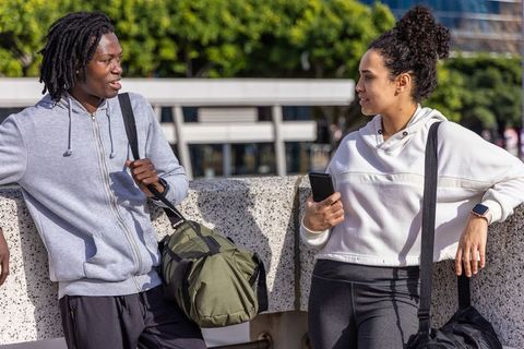 Young diverse friends chatting after workout leaning on concrete barrier with duffel bags