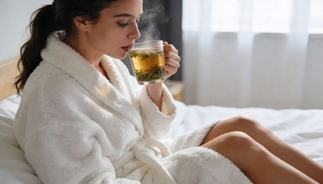Woman relaxing in plush white bathrobe sipping steaming herbal tea on cozy morning bed