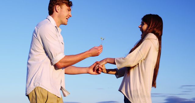A young Caucasian man offers a flower to a young Caucasian woman, both smiling and engaging in a romantic gesture, with copy space. Their interaction suggests a moment of affection and connection during a beautiful time of day.