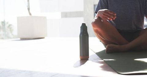 Man meditating on yoga mat beside insulated bottle in sunlit minimalist interior