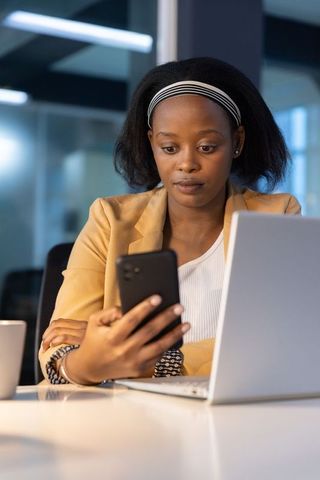African american woman checking smartphone while working at laptop in modern office