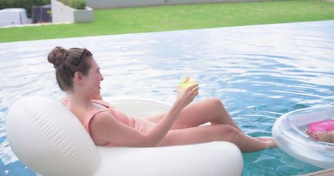Woman reclining on white inflatable pool lounger holding yellow drink relaxing in sunlit clear pool