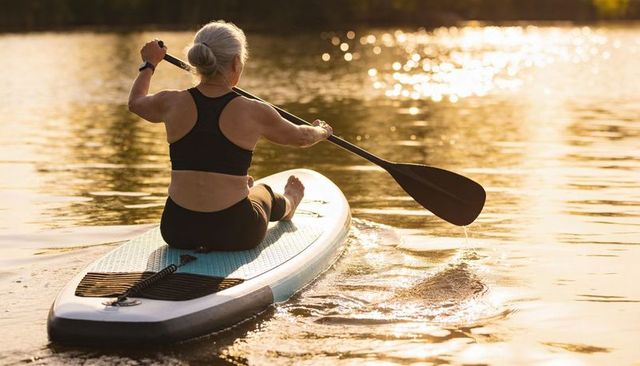 Senior woman paddling stand up paddleboard during golden sunrise on calm lake