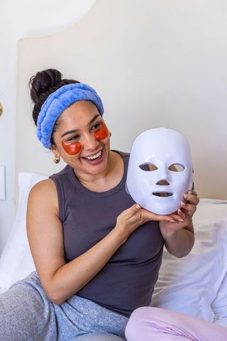 Smiling woman holding led facial mask while wearing under-eye gel patches and headband