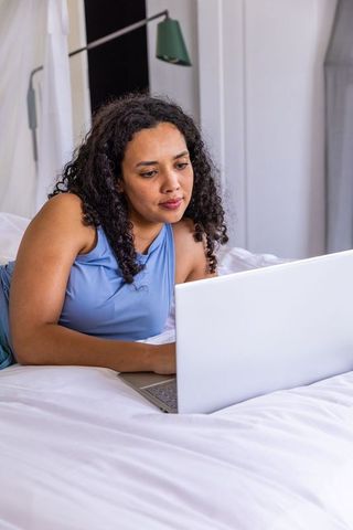 African american woman reclining on bed using laptop, wearing sleeveless light blue top