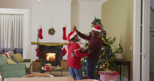 Couple wearing Santa hats dancing joyfully near fireplace decorated for Christmas. Cozy living room filled with festive decorations and Christmas tree in background. Suitable for holiday season campaigns, family festivity themes, or advertising warm and festive environments.