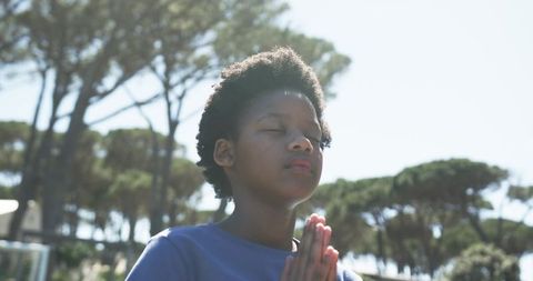 Sunlit serene african american teen praying with hands pressed together in pine tree park