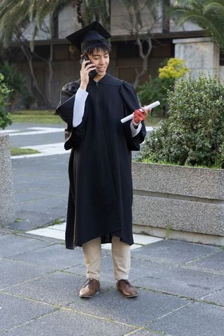 Asian graduate celebrating on campus holding diploma and calling family in cap and gown
